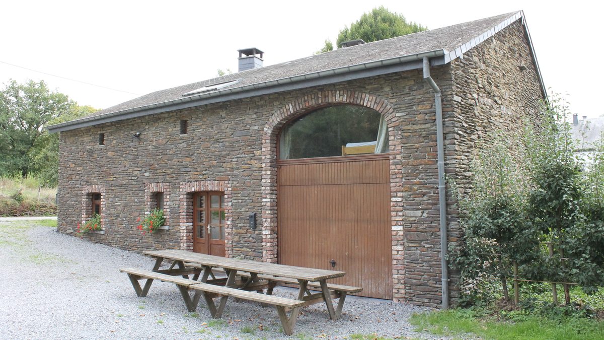 De natuurstenen hoeve van opzij gezien, met de grote boogpoort en twee picknicktafels op het grind ervoor.